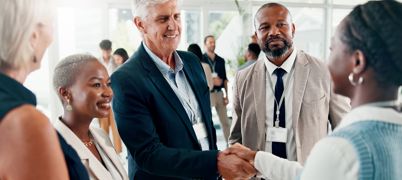 Grey haired man smiles and shakes hand with woman with two people looking on