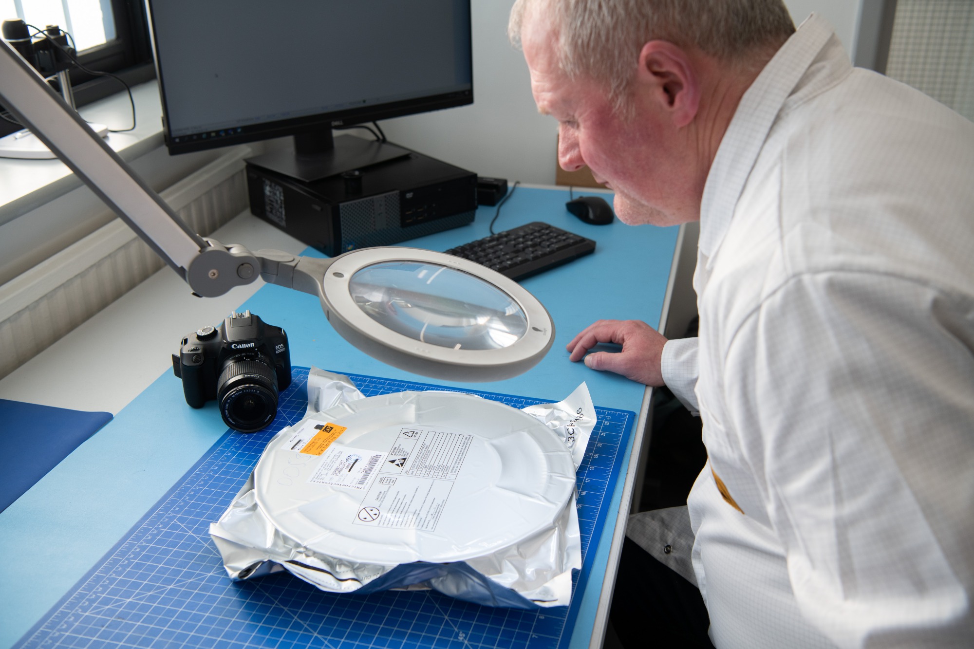 A male IC Blue member of staff, inspecting an electronic manufacturing component at his desk
