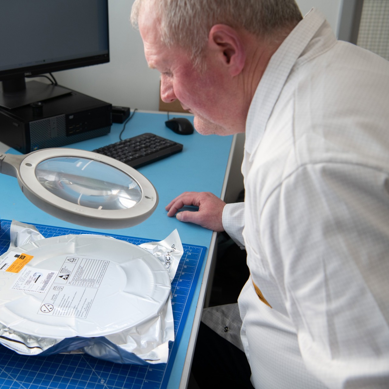 A male IC Blue member of staff, inspecting an electronic manufacturing component at his desk