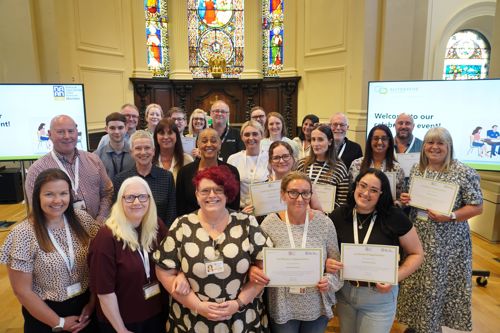 A group of people standing indoors in three rows with some holding certificates with a church backdrop