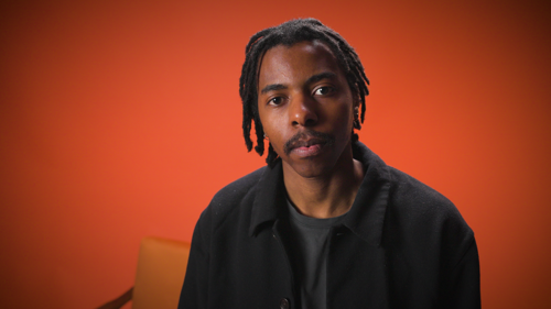 Local artist Nigel looking into the camera with a seat behind him with an orange background