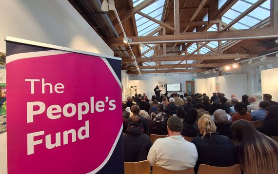 Pink and blue roll up banner says 'The People's Fund' in the foreground behind it is a seated crowd listening to a presentation
