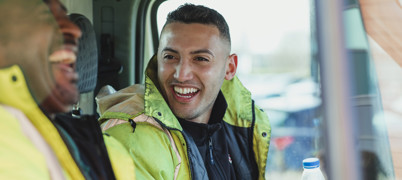 Two Male Labourers Wearing High Vis Jackets Talking And Laughing In Their Work Van Together