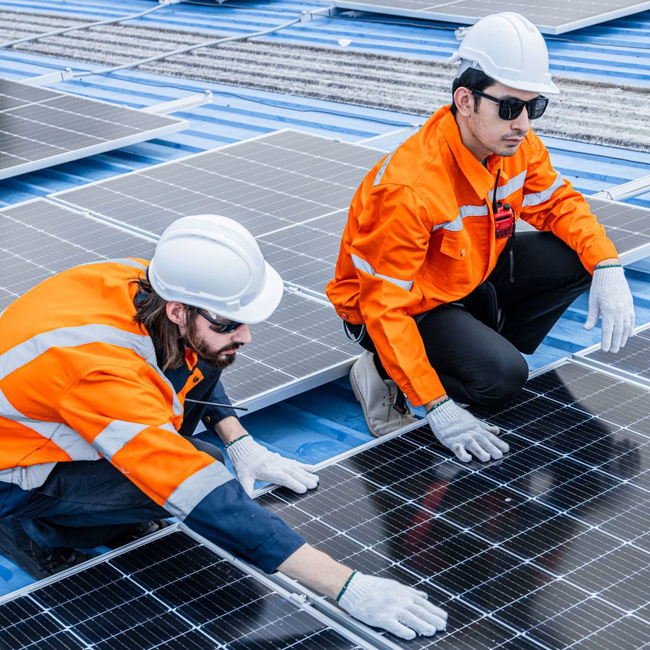 Two men in hard hats and orange jackets work on solar panels