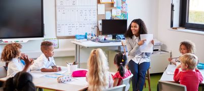 Female primary school teacher sitting on a chair facing school children in a classroom holding up and explaining a worksheet to them