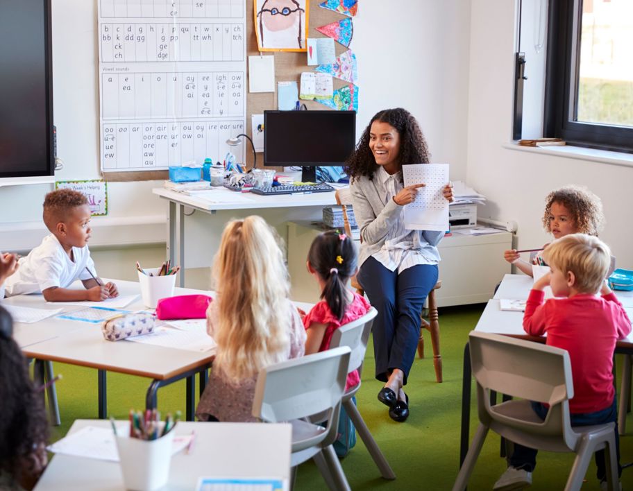 Female primary school teacher sitting on a chair facing school children in a classroom holding up and explaining a worksheet to them