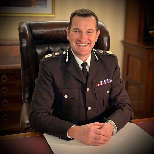 Sir John Robins in uniform smiling sitting at desk in a brown chair with his hands on the table in front of him