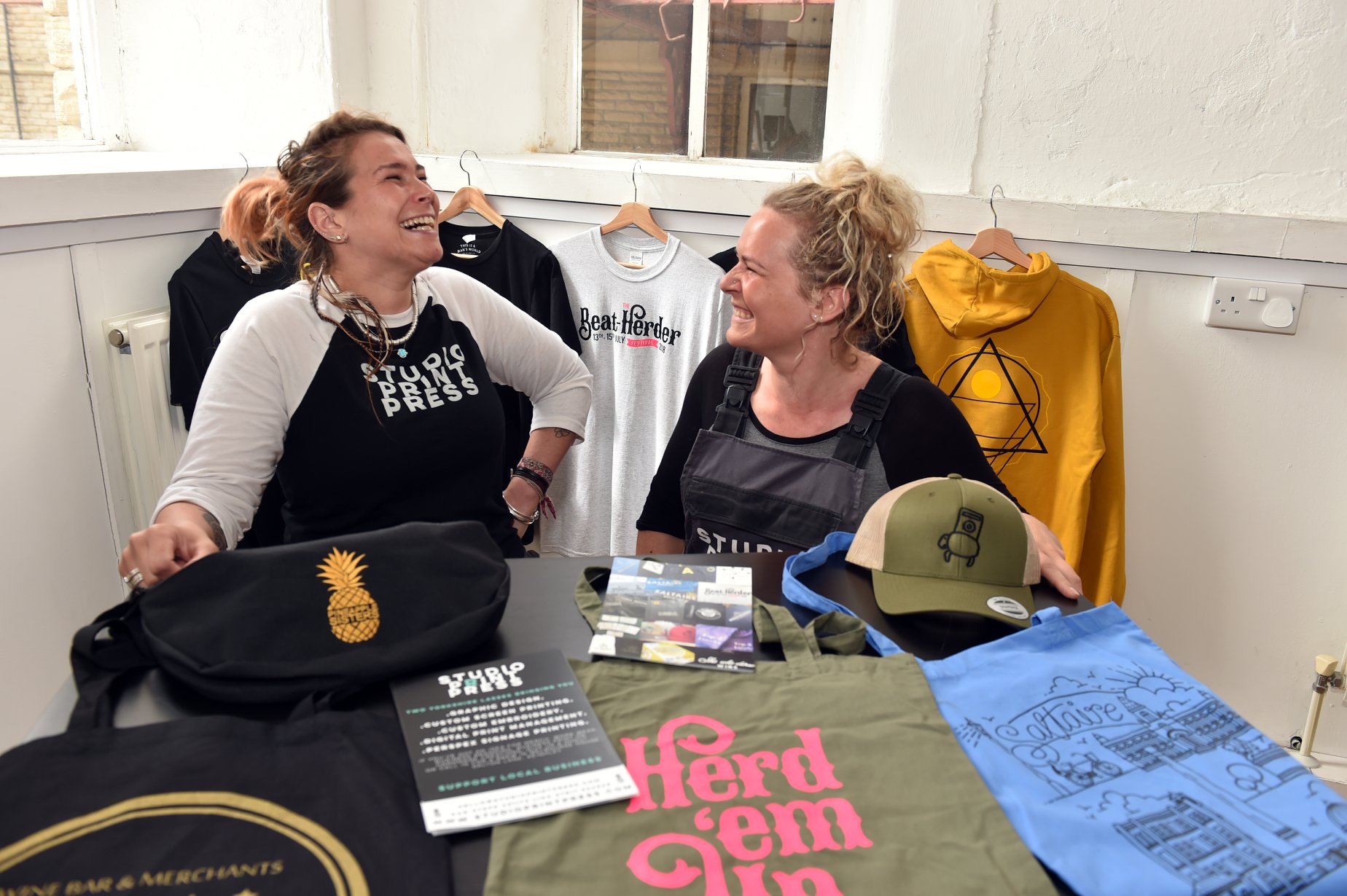 Two ladies laughing with a range of printed merchandise in front of them